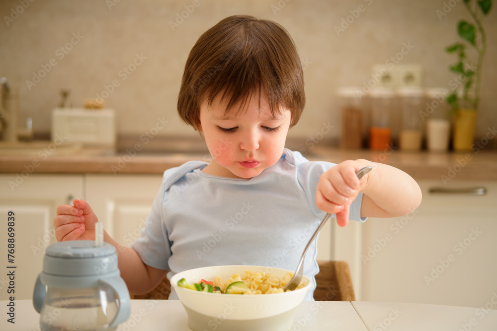 A child eats vegetable salad with a fork while sitting at a table in a home kitchen. Cute little boy eating pasta. Toddler baby two years old at dinner A child eats vegetable salad with a fork while sitting at a table in a home kitchen. Cute little boy eating pasta. Toddler baby two years old at dinner