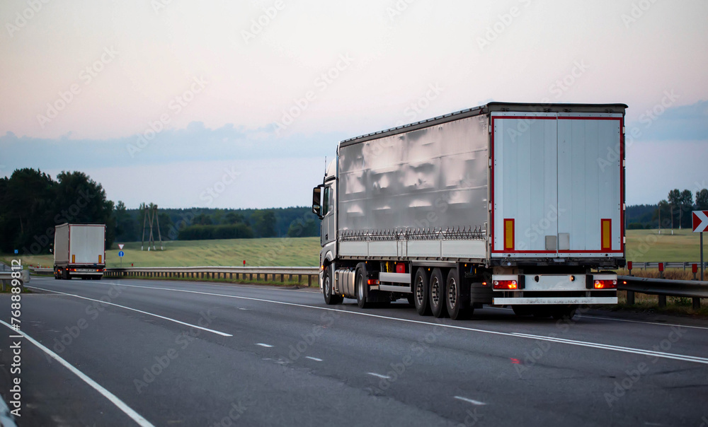 A column of new trucks with tilt semi-trailers drives along a country road in the evening in the summer against the backdrop of a forest. Concept of cargo transportation by logistics. 