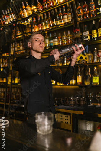 Bartender shakes cocktail for guest looking straight standing by bar counter with glass of ice. Barman of Ukrainian pub fulfills order
