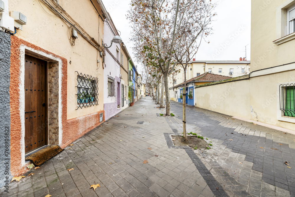 Facades of old houses of various colors of two heights with metal bars on the windows and trees with tree pits on a pedestrian street with conventional sidewalks Facades of old houses of various colors of two heights with metal bars on the windows and trees with tree pits on a pedestrian street with conventional sidewalks