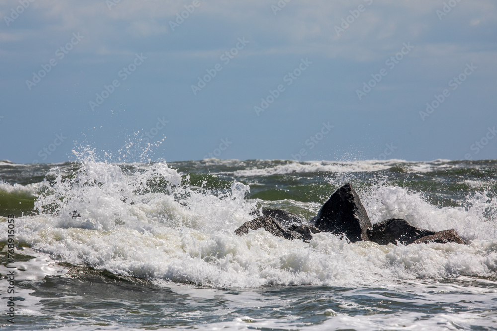 Fototapeta premium Waves crash on dark rocks with a uniform blue sky above. White Sea foam and ocean spray and small waves in the foreground. Waves crash during high tide in Florida on the Atlantic Ocean coastline.