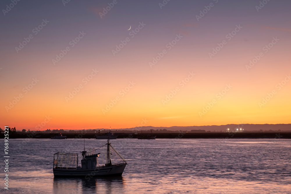 Sunset prices on a Spanish beach with beautiful clouds, calm sea and a fishing boat in the foreground and beautiful concave crescent moon.