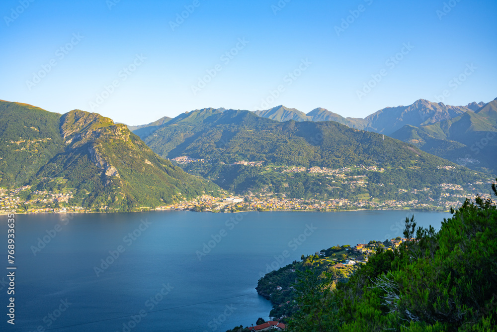 A beautiful mountain range with a lake in the foreground. The lake is calm and the mountains are covered in trees. The sky is clear and the sun is shining brightly. The scene is peaceful and serene