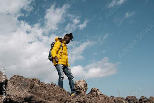 Canvas-taulu Young African man with vibrant yellow jacket - hiking rocky landscape, adventure fashion - clear sky, outdoor exploration - copy space