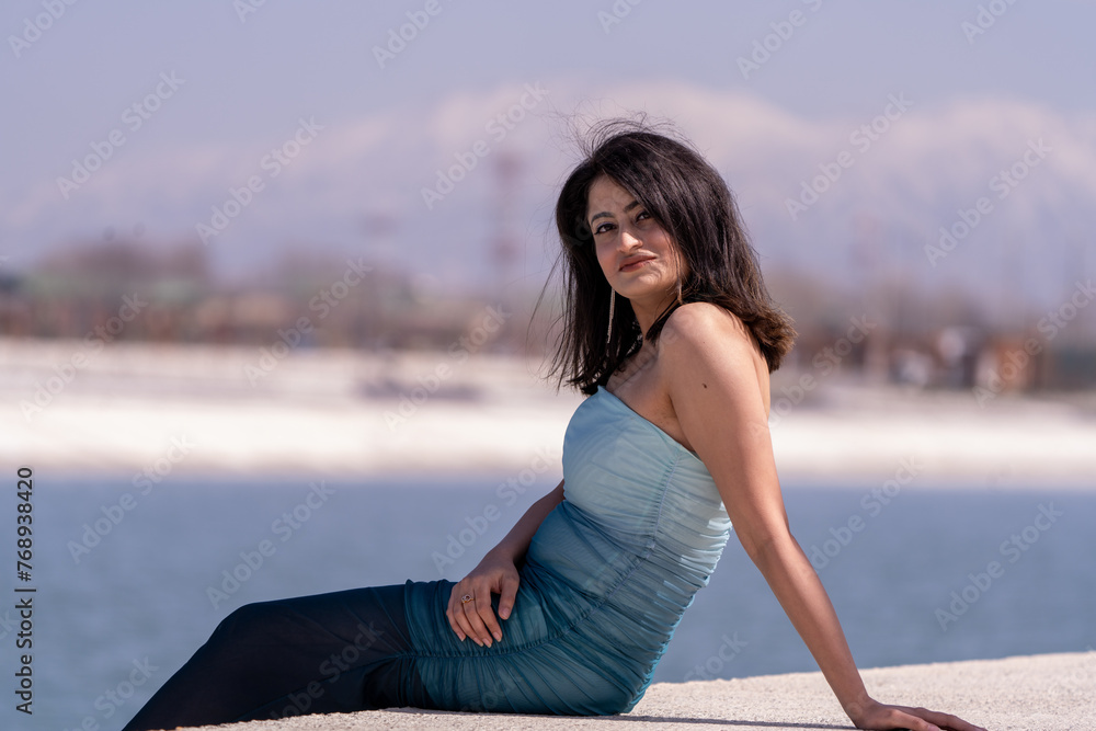 A woman is sitting on a ledge by the water, wearing a blue dress