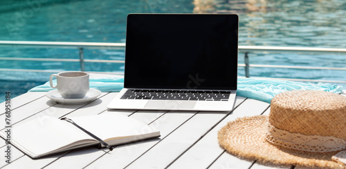 Alternative workplace at resort poolside. Laptop, notepad, cup of coffee and hat on white table isolated on blue background of pool water. Blank computer screen, copy space for text or design. Banner