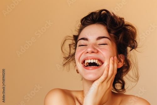 woman laughing with her hands, her head tilted back and eyes closed in joy. She has short hair styled. The background is neutral with soft lighting, highlighting her expression and skin texture. © zayatssv