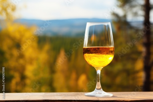 A stem glass of golden amber wine on a wooden surface with a blurred autumnal forest backdrop. Glass of Amber Wine Overlooking Autumn Forest