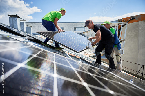 Workers building solar panel system on metal rooftop of house with assistance of crane lift. Two men installers carrying photovoltaic solar module outdoors. Renewable energy generation concept.