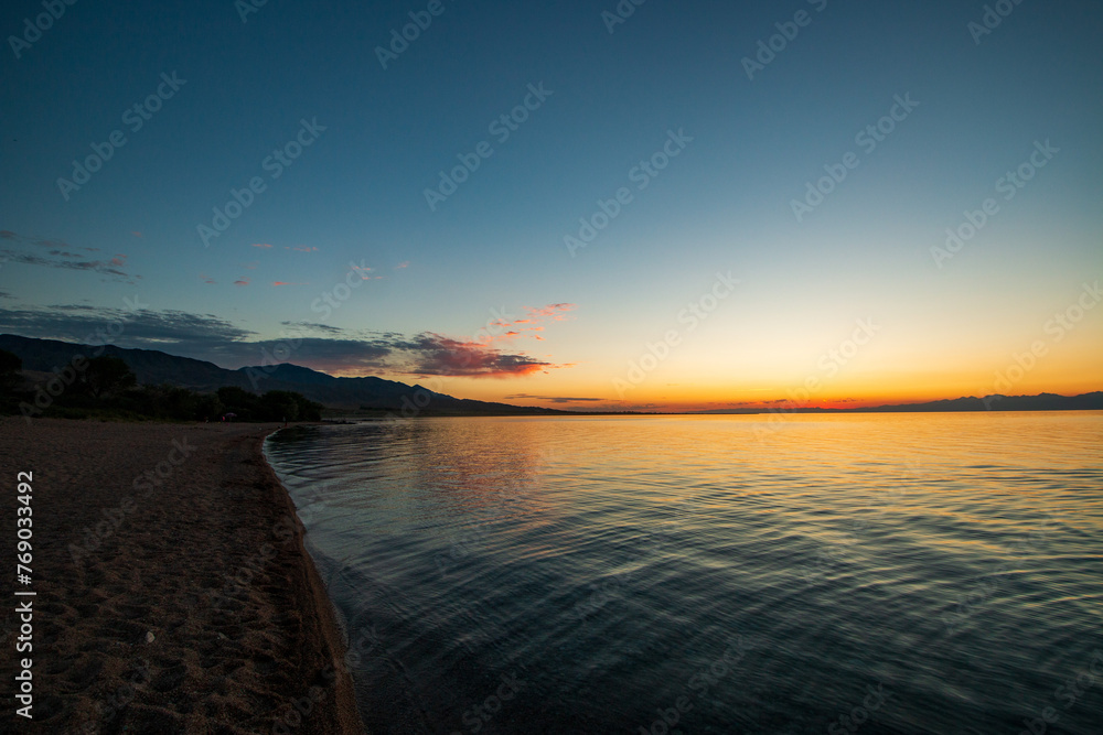 Fototapeta premium An old man walking by the shore of the famous Issyk-Kul lake surrounded by the Ala-Too mountains. Kyrgyzstan, July 2023