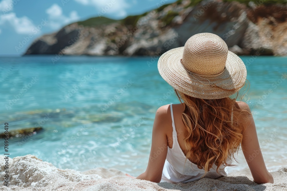 An inviting view of a woman in a white dress seated on sand, gazing into the clear turquoise sea and rocky landscape