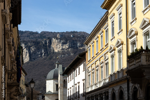 View of buildings and the Cathedral Tower in a sunny day in Trento city centre. Dolomites in background; Trentino-Alto Adige, Italy