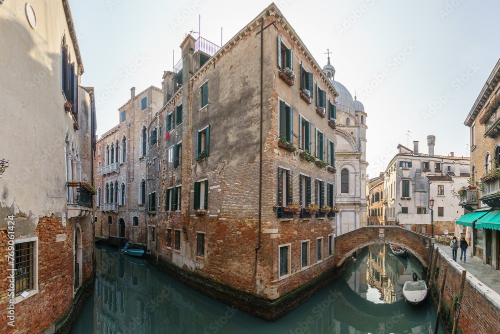 Typical narrow canal surrounded by buildings with boats and a footpath bridge in Venice, Veneto, Italy