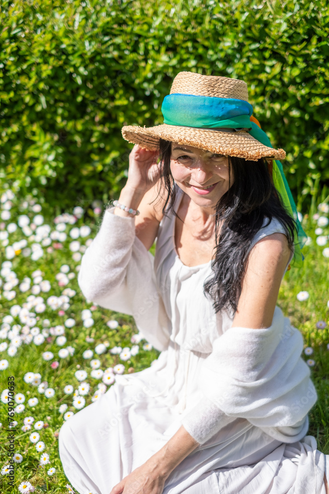 Smiling woman in white, straw hat with blue ribbon, amidst a green and white daisy field, joyful summer vibe, nature embrace, sunny backdrop.