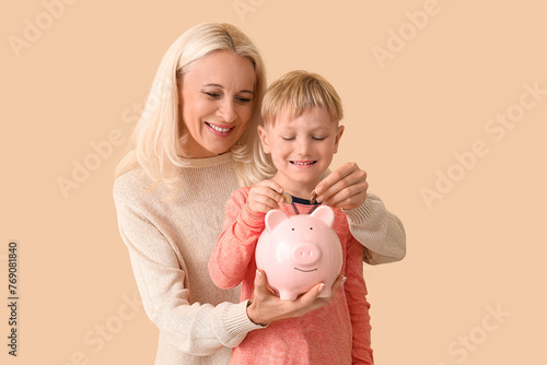 Obraz na płótnie Little boy with his grandmother putting coins in piggy bank on beige background