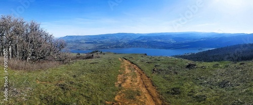View looking down a mountain biking trail on a steeply sloping meadow with a few small pine trees down near a massive river winding out of view. 