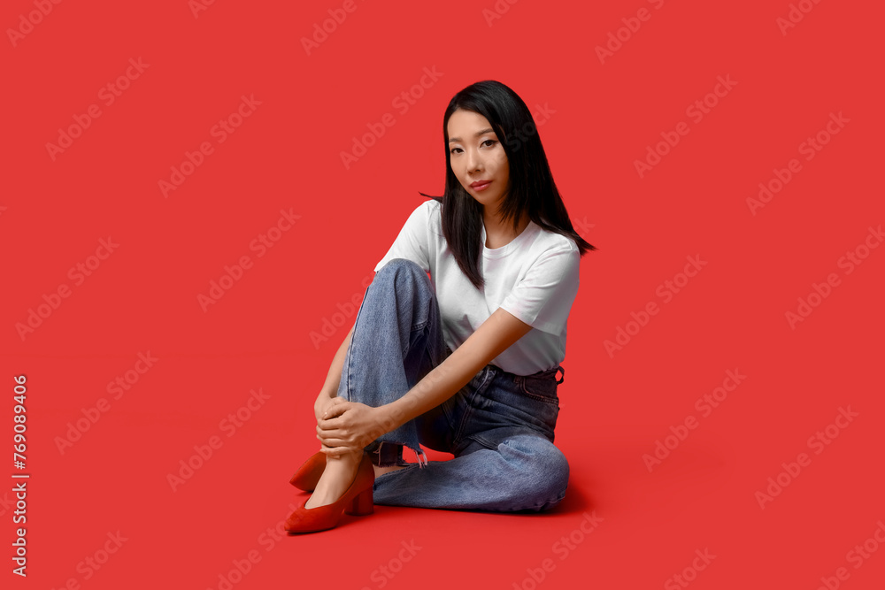 Beautiful young Asian woman in stylish jeans sitting on red background Beautiful young Asian woman in stylish jeans sitting on red background