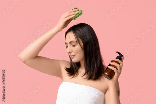 Beautiful young woman with hair scalp massager and bottle of shampoo on pink background photo