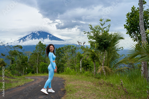 A young woman in a blue tracksuit walks along a road in the mountains. Hiking with a view of the popular holy volcano Agung covered in clouds on the island of Bali.