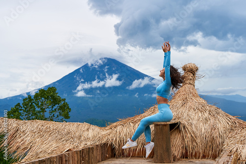 A young woman poses on a large straw photo zone as a bird on a viewing platform and enjoys the view of the sacred Mount Agung volcano hidden by clouds on a rainy day on the island of Bali.