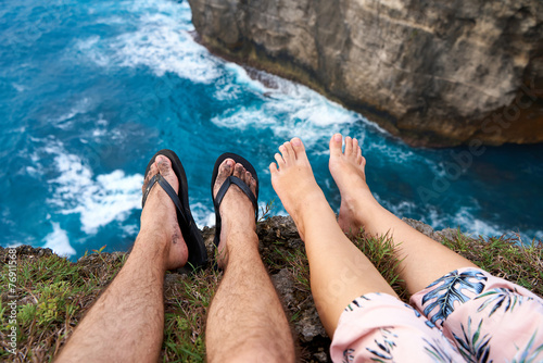 A couple sits on a cliff with their feet dangling over an abyss above the ocean. First person photo.