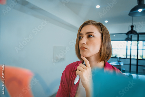Fototapeta Naklejka Na Ścianę i Meble -  Portrait of young caucasian businesswoman thinking with confused face expression while standing in front of glass board with sticky notes and mind map at creative business meeting. Immaculate.