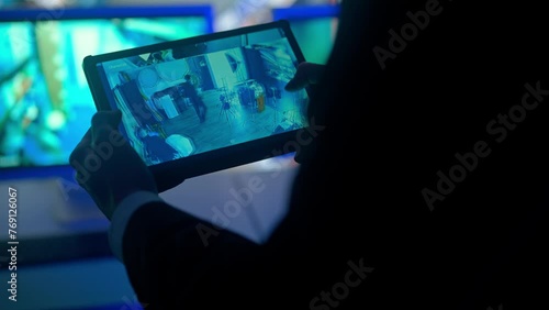Man security guard officer working on tablet in monitoring center, zooming watching video. Computers and big digital screen show cctv cameras footage.