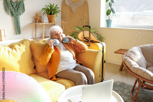 Senior female tourist with neck pillow sitting on sofa at home
