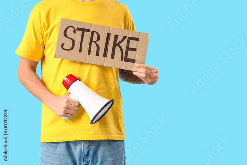 Protesting young man holding placard with word STRIKE and megaphone on blue background, closeup © Pixel-Shot