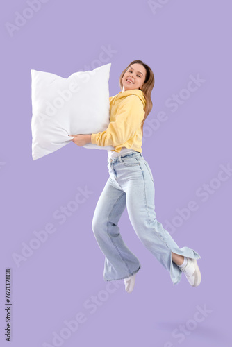 Young woman with soft pillow jumping on lilac background