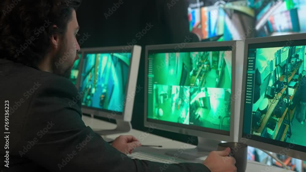 Man security guard officer working in monitoring center, watching cameras, drinks coffee. side shot. Computers and big digital screen show cctv cameras footage. Man security guard officer working in monitoring center, watching cameras, drinks coffee. side shot. Computers and big digital screen show cctv cameras footage.