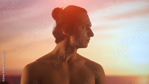 Profile of a young man with bare torso admiring a sunset, his face illuminated by the warm golden light of the twilight sky.