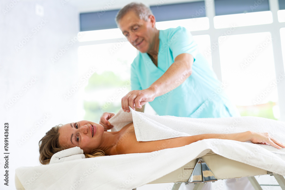Obraz premium Relaxed middle-aged woman lying on massage table covered with soft blanket by kind masseuse