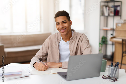 Happy black male student writing notes with laptop