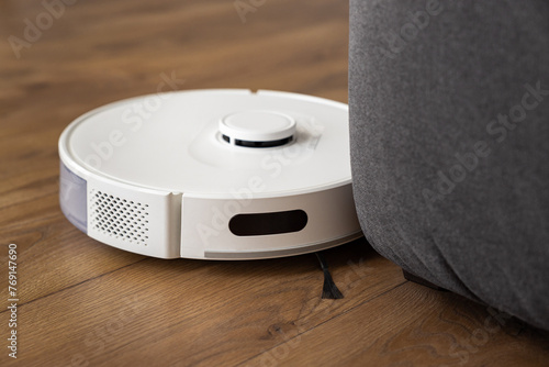 A white robot vacuum cleaner cleans a dark oak wood laminate on the floor. collects dust near the sofa in difficult places. automatic management of modern technologies. infrared sensors control