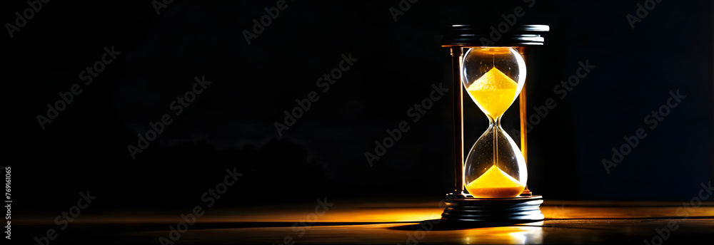 A sand timer placed on a wooden table in the dark with dim lighting. Yellow sand inside the timer indicates that time is running out - obrazy, fototapety, plakaty 