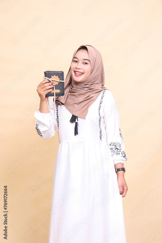 Fototapeta premium Beautiful Asian Muslim adult woman wearing a hijab with a cheerful expression tilted carrying prayer beads tied to the Koran while holding a dress. for advertising, lifestyle, banners and Ramadan