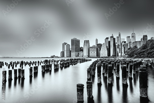 Black and white photo of the iconic manhattan skyline over calm waters, with weathered pier remains in the foreground