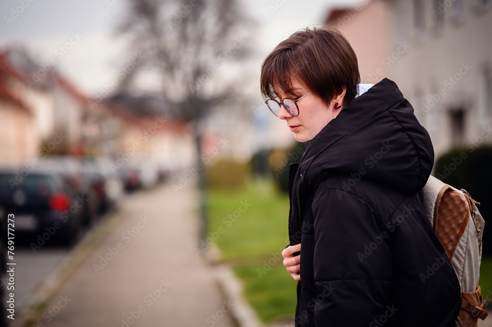 A young adult woman, dressed warmly in a black jacket with a hood and glasses, gazes thoughtfully off-camera in an outdoor, suburban setting