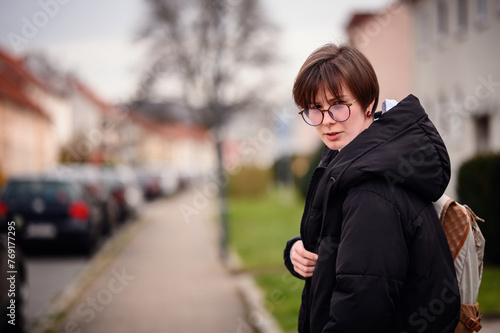 A young adult woman in casual attire, with a stylish haircut and round glasses, stands on a suburban street, carrying a backpack over one shoulder.