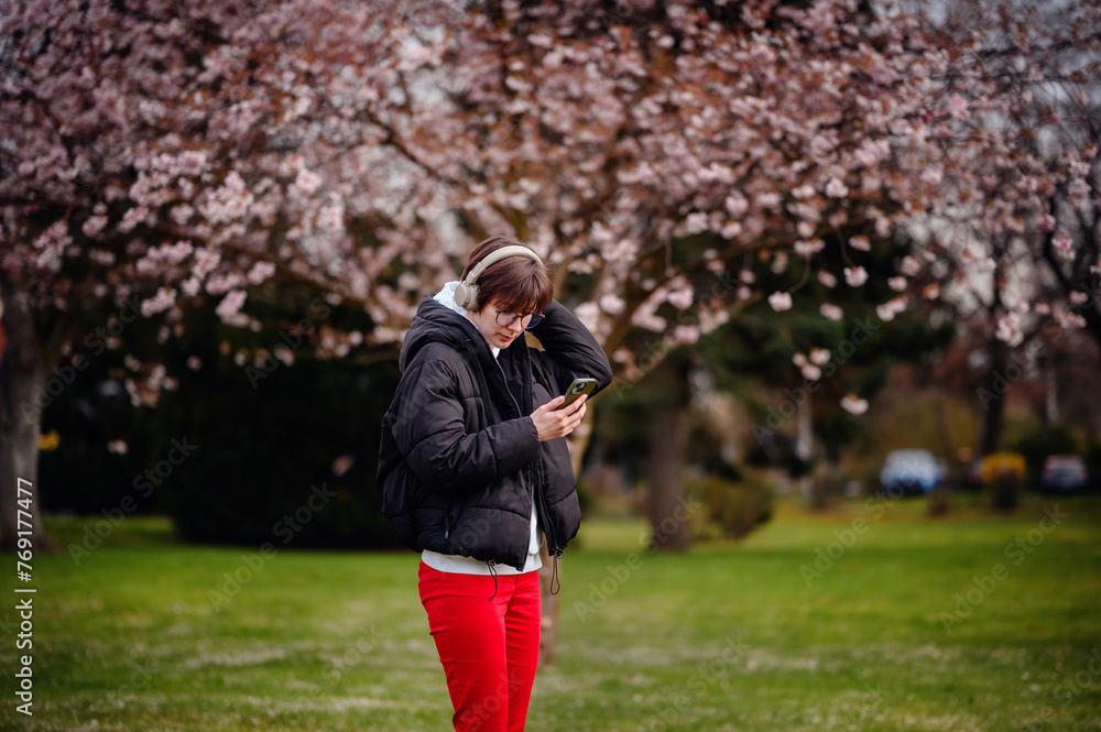 Engrossed in her smartphone and music, a young woman stands under the spring bloom of cherry blossoms, her headphones suggesting a moment of leisure and relaxation Engrossed in her smartphone and music, a young woman stands under the spring bloom of cherry blossoms, her headphones suggesting a moment of leisure and relaxation