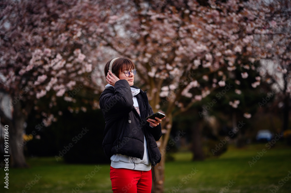 A woman with a contented smile enjoys the melody of her music amidst the delicate pink of cherry blossoms in a serene park setting A woman with a contented smile enjoys the melody of her music amidst the delicate pink of cherry blossoms in a serene park setting