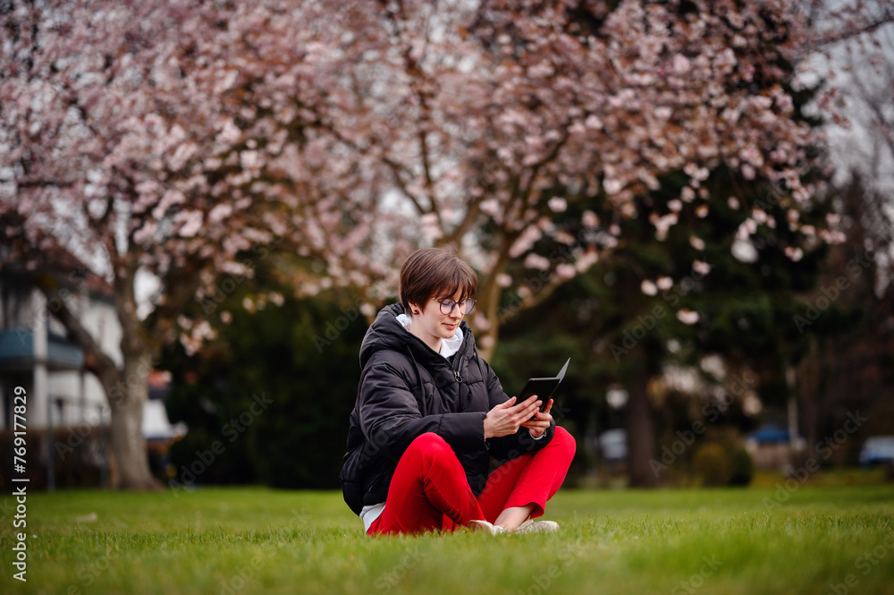Fototapeta premium A focused young woman sits cross-legged on the grass in a park, deeply engaged with her tablet with a beautiful cherry blossom tree flowering in the background