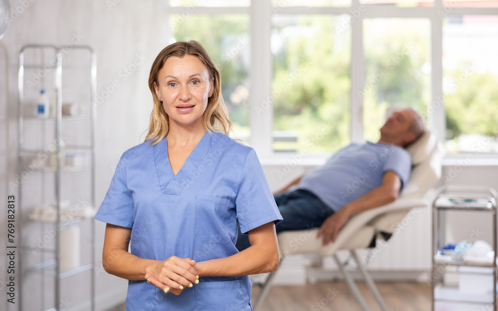 Fototapeta premium Portrait of confident professional female doctor in blue scrub standing in medical office on blurred background with patient lying on couch