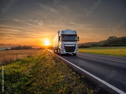 Fototapeta Naklejka Na Ścianę i Meble -  White truck driving on the asphalt road in rural landscape at sunset
