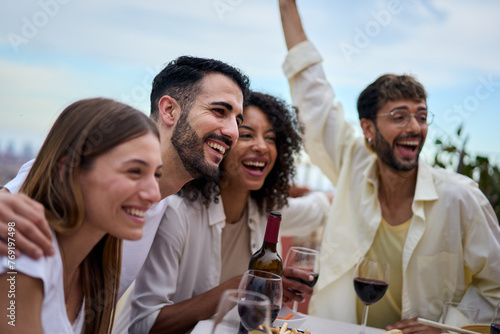 A group of happy humans sitting at a table, with smiles and wine glasses, enjoying leisure time together. The sky above them as they gesture and have fun in a community setting