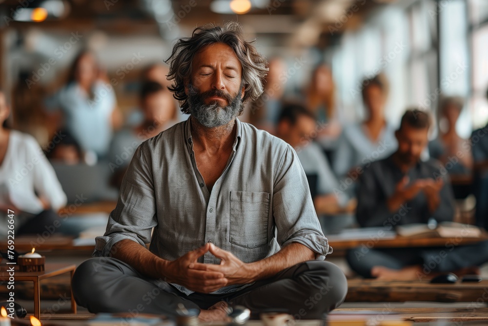 A man with a beard and moustache is sitting in a lotus position with his eyes closed, participating in a meditation event in front of a group - obrazy, fototapety, plakaty 