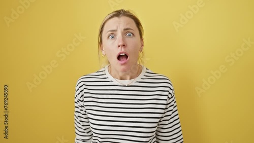 Attractive young blonde woman, skeptical look on her uncertain face, sarcastically standing in disbelief with open mouth against an isolated yellow background