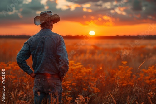 A contemplative man in a cowboy hat stands overlooking a golden country field at sunset