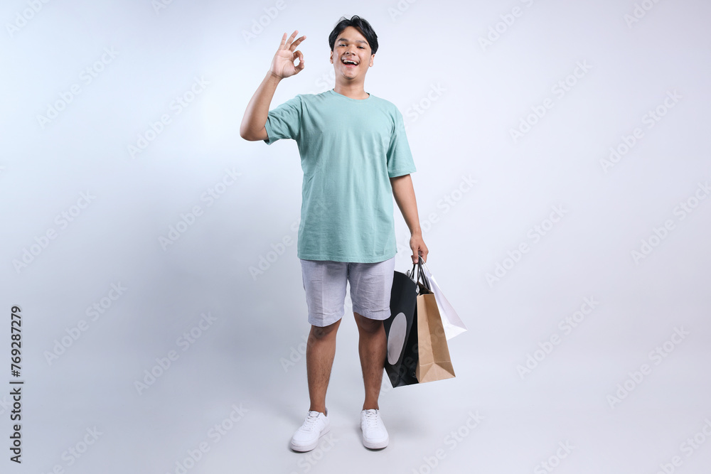 Full length view of young Asian man wear casual summer clothes showing ok sign with finger while holding shopping bags Full length view of young Asian man wear casual summer clothes showing ok sign with finger while holding shopping bags